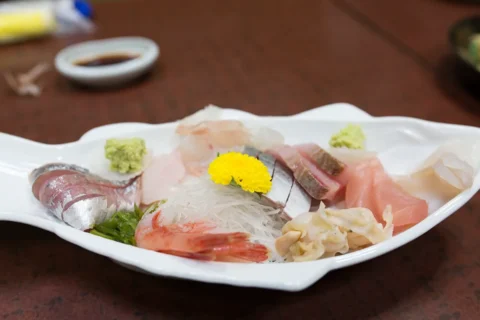 白い舟形の皿に、青魚・甘えび・赤身などの刺身が並び、中央の大根のけんの上に黄色い食用菊、手前に緑の海藻、両端にわさびが添えてある