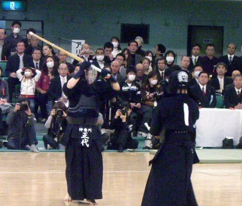 剣道の試合で、選手が竹刀を頭の上に高く振りかぶって構え、相手と向かい合っている