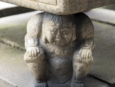 石の下にかがみこんで重みを支える天邪鬼の石像